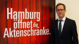 Projektleiter André Basten steht vor einem Werbeplakat der Stadt Hamburg mit der Aufschrift "Hamburg öffnet die Aktenschränke".
